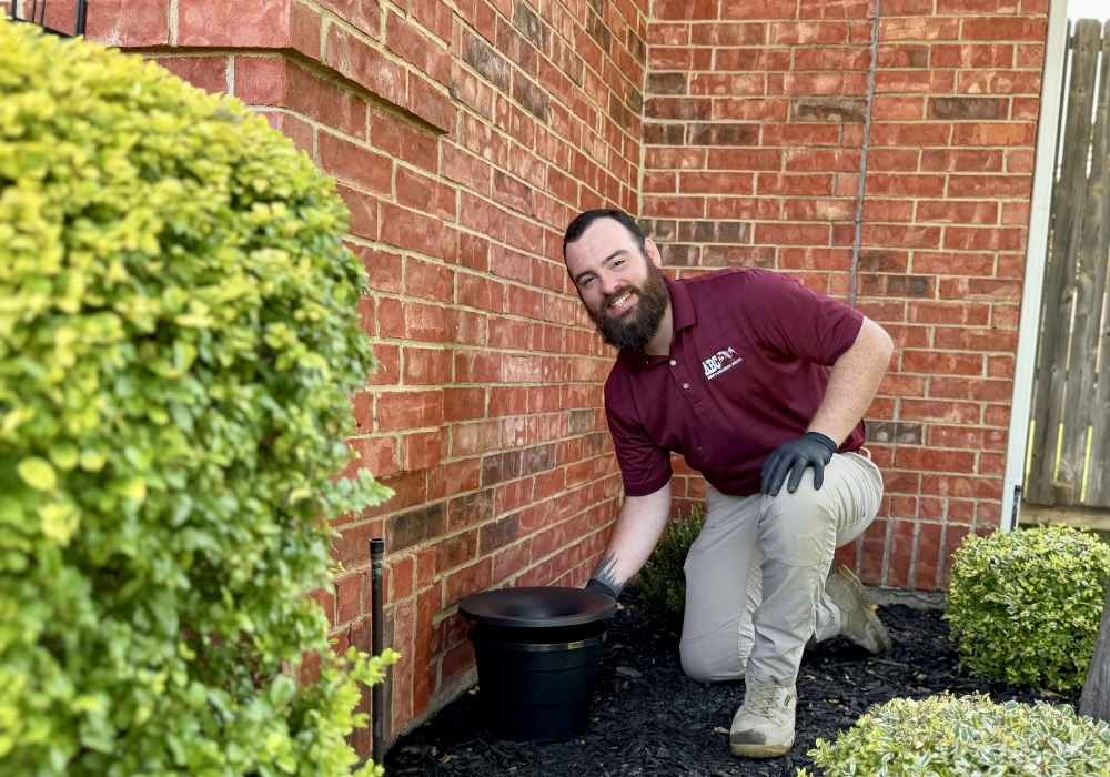 ABC Home & Commercial Services technician installing a mosquito misting system along a brick home’s foundation in a landscaped yard.